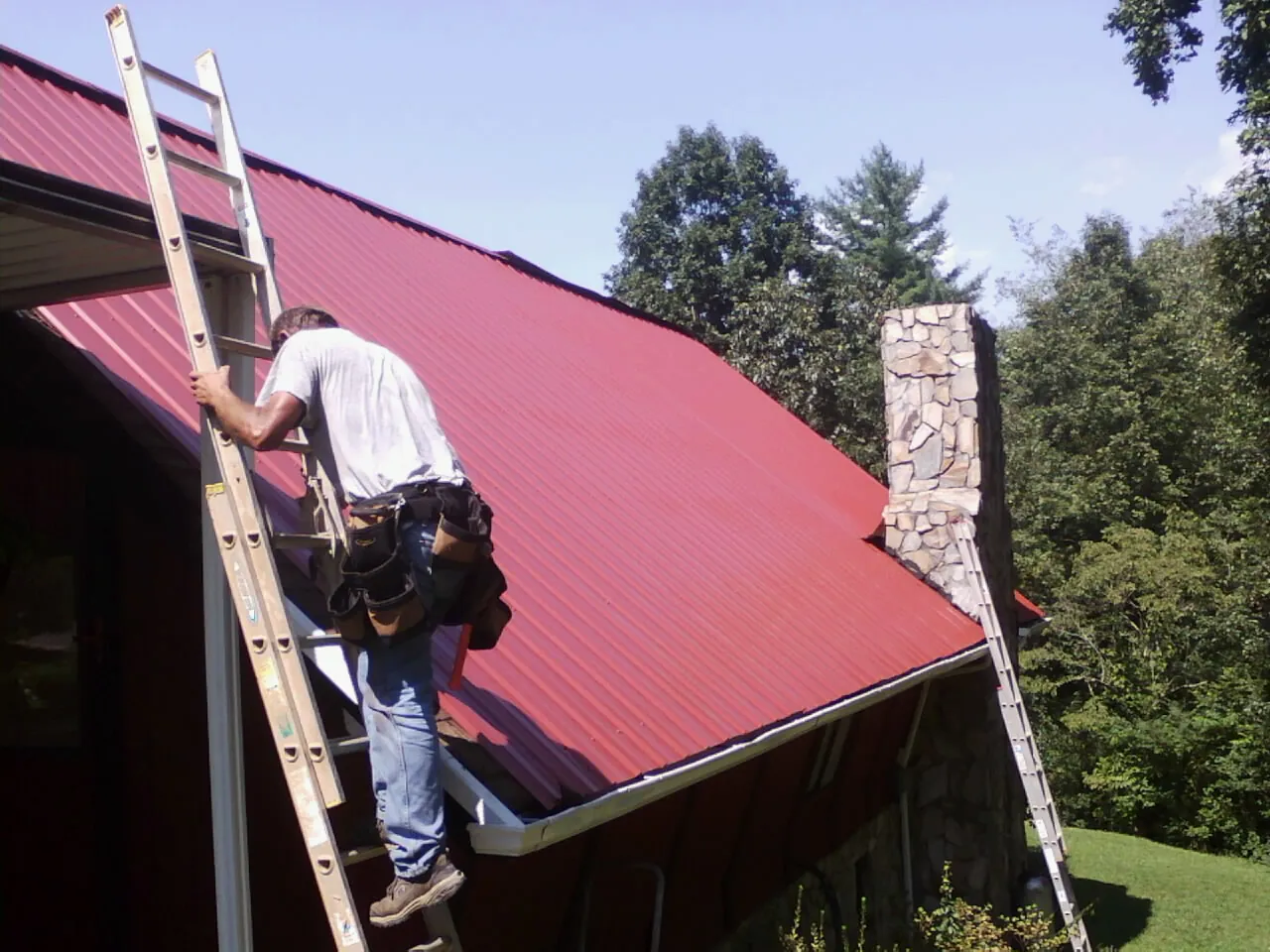 Roofer on a ladder beside a red metal roof and stone chimney — Soffit & Fascia Repair by Roofing Brothers in Avon